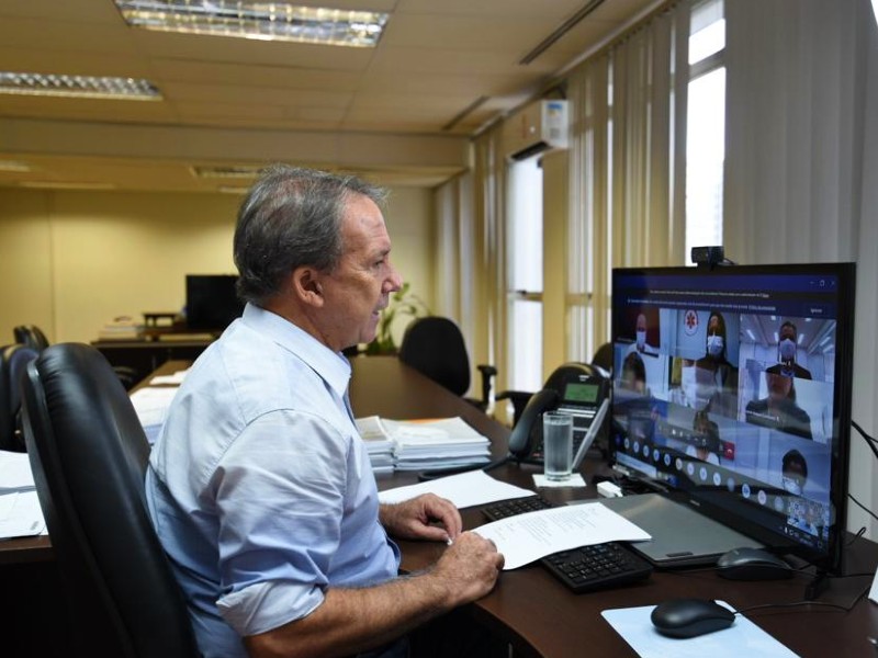 Foto do secretário da Saúde, Edson Aparecido. Ele está em seu gabinete, sentado numa cadeira e de frente para uma tela onde acompanha a cerimônia virtual da entrega do Prêmio Cidade de São Paulo